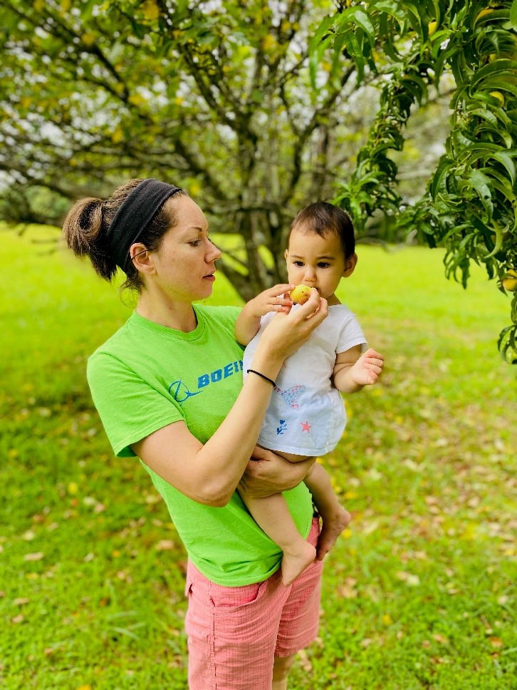Romina is standing in her orchard with Zara in her arms. She is feeding Zara the peach.